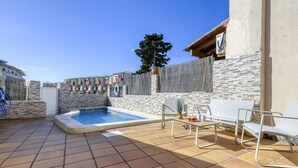 Una piscina con un jacuzzi, rodeada de muros de piedra y una valla de madera. Hay sillones blancos para tomar el sol y una mesa con un frutero.