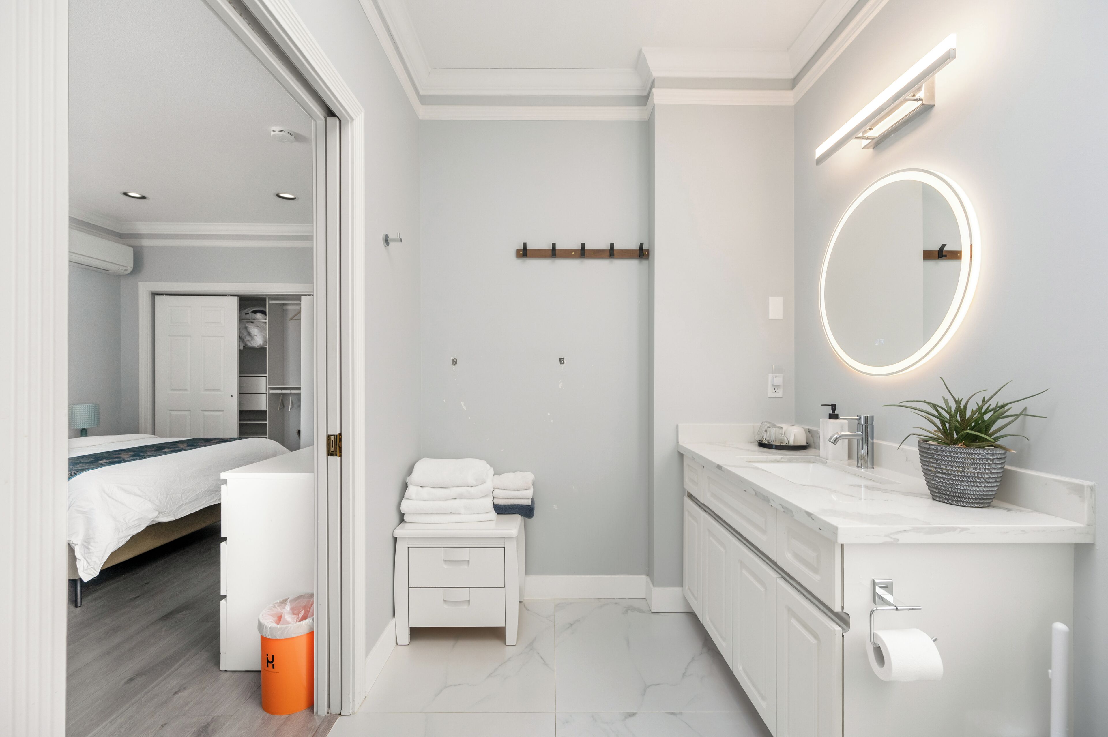 Et moderne badeværelse med et hvidt underskab, et rundt spejl med lys, en potteplante og en toiletpapirholder.