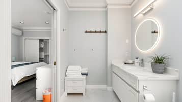 Et moderne badeværelse med et hvidt underskab, et rundt spejl med lys, en potteplante og en toiletpapirholder.