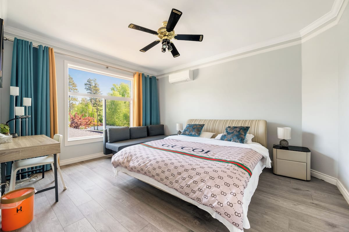 A bedroom with a bed, a desk, a chair, a nightstand, a lamp, a ceiling fan, and a window with curtains.