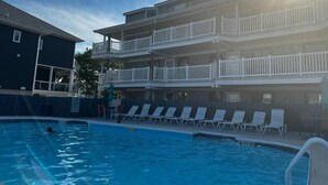 Una piscina con sillones de descanso y un edificio de varios pisos con balcones.