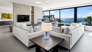 A modern living room with a large sofa, a coffee table, and a TV mounted on the wall. There is a large window with a view of the ocean.