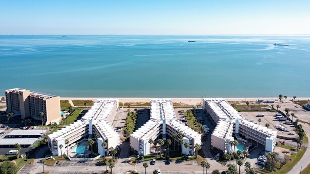 A hotel complex with multiple buildings, a pool, and a beach view. - Just Beachy (Corpus Christi)
