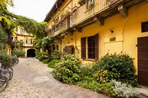 A narrow cobblestone path leading to a yellow building with balconies, surrounded by lush greenery and bicycles.