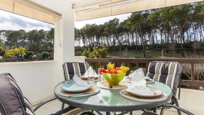 A round glass table with striped cushions, a bowl of fruit, and a glass of water on a balcony with a view of trees and a fence.