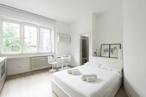 Una habitación de hotel moderna con una cama grande, un escritorio, una silla y un ventanal con vistas a edificios y árboles.