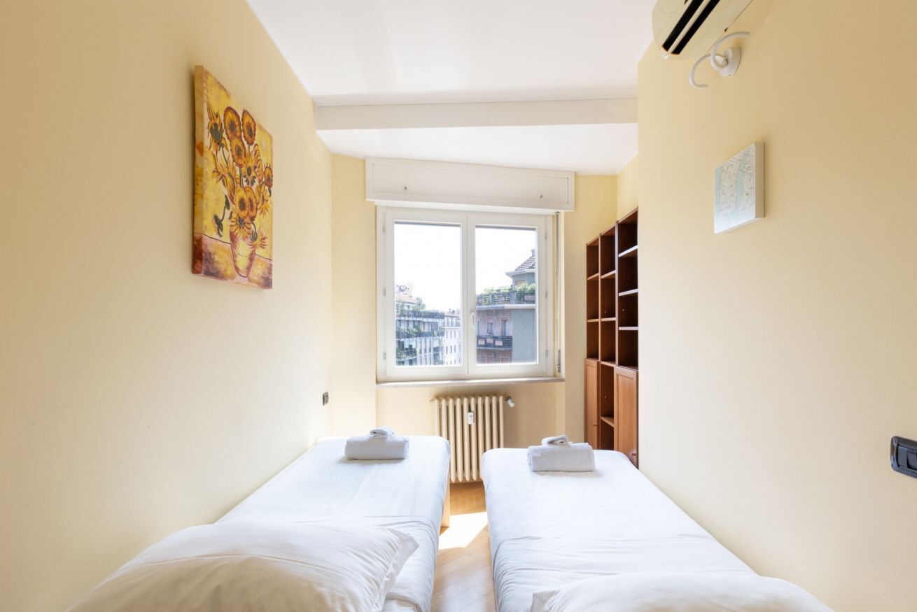 A room with two beds, a window with a view of buildings, a radiator, and a shelf with books.