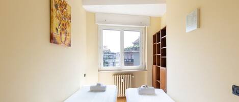 A room with two beds, a window with a view of buildings, a radiator, and a shelf with books.