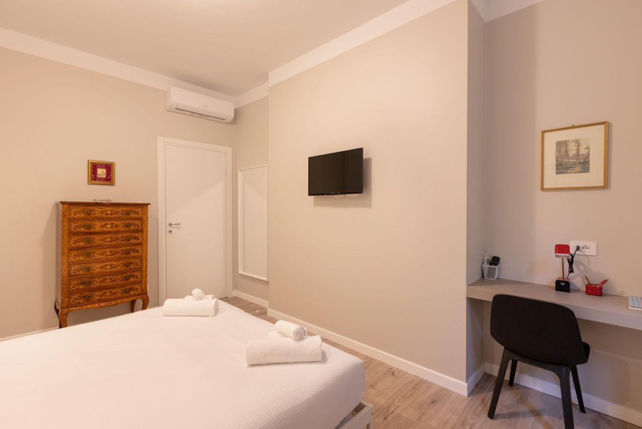 A bedroom with a bed, a wooden dresser, a desk with a chair, a flat-screen TV, and a framed picture on the wall.