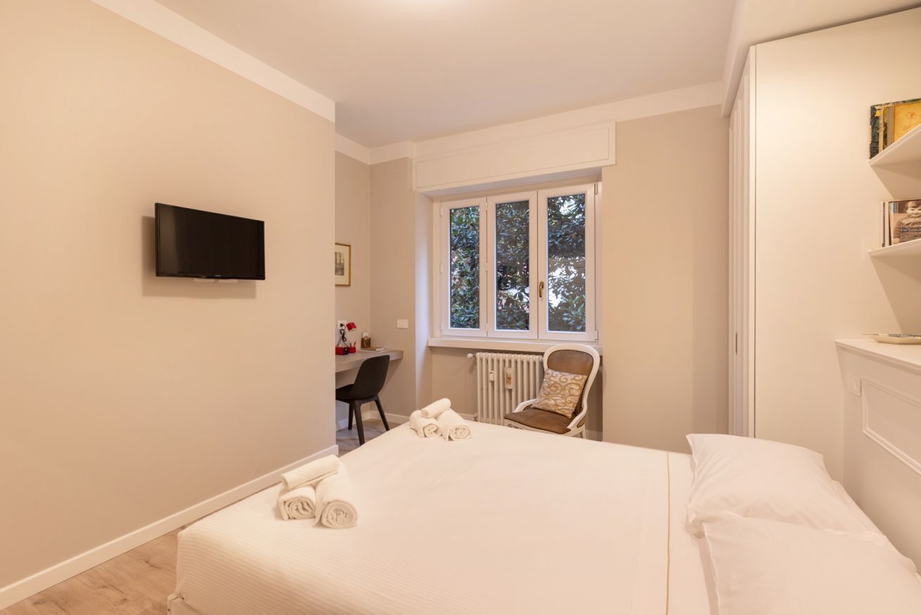 A bedroom with a bed, a desk, a chair, a television, and a window.