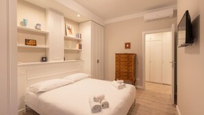 Quarto de hotel com cama, paredes brancas, cômoda de madeira, televisão e guarda-roupa.