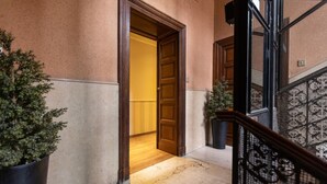 Un pasillo con una puerta de madera que da a una habitación, una escalera con barandilla de hierro forjado y plantas en macetas.
