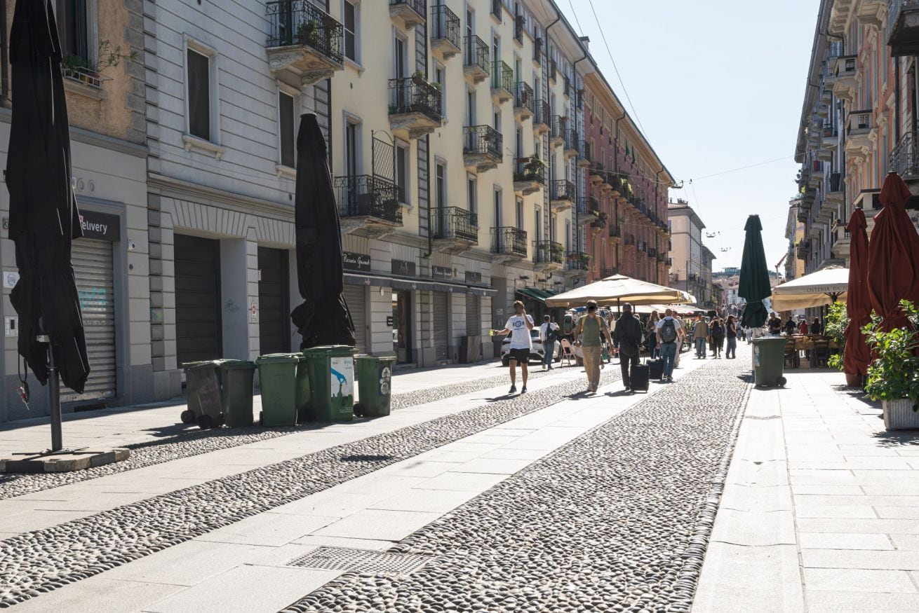 Calle peatonal flanqueada por edificios, con mesas al aire libre y contenedores de basura.