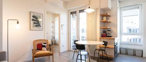 Una sala de estar moderna con una mesa redonda, dos sillas, un sofá y un sillón de mimbre. Se ve un ventanal con persianas, una lámpara de techo y un cuadro en la pared.