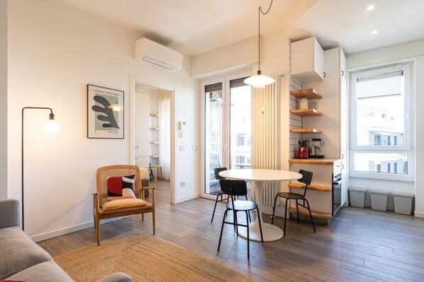 A modern living room with a round table, two chairs, a sofa, and a wicker chair. There is a large window with blinds, a ceiling light, and a framed picture on the wall. - Modern one bedroom Navigli - Mirable PM (Milano)
