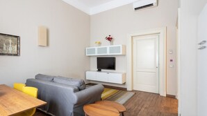 A modern living room with a grey sofa, a wooden dining table with yellow chairs, a white entertainment unit with a TV, and a door leading to another room.