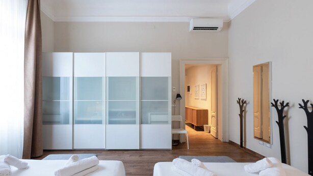 A hotel room with two beds, a wardrobe with frosted glass doors, a bathroom visible through an open door, and a hallway with a mirror and wall-mounted decorations.