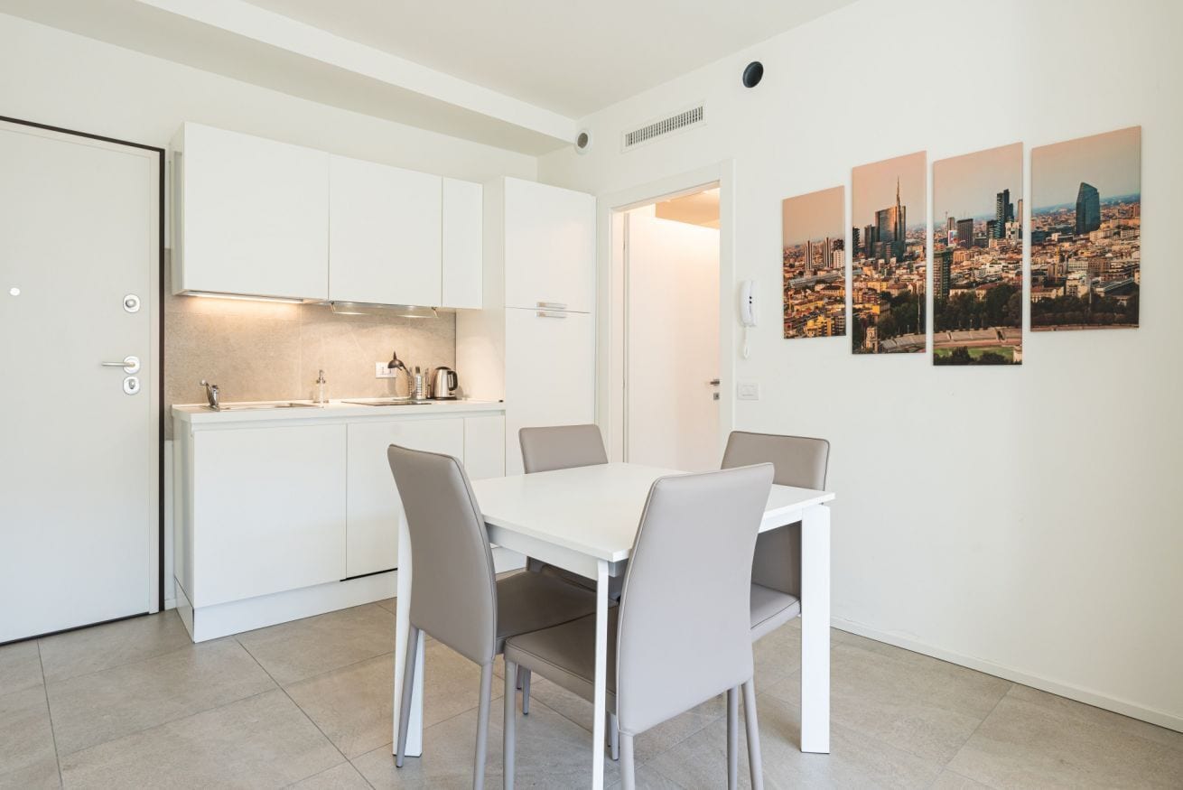 Una cucina moderna con mobili bianchi e un'area pranzo con un tavolo bianco e quattro sedie. Alle pareti, quadri raffiguranti una vista cittadina.