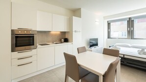 A modern kitchen with white cabinets, a built-in oven, and a sink. There is a dining table with chairs and a bed with white linens.