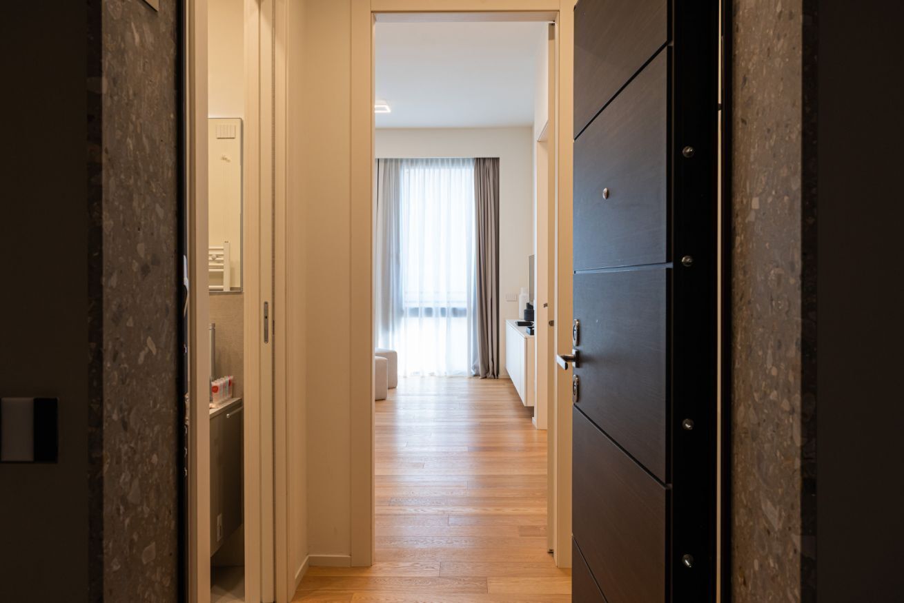 Un pasillo con suelo de madera, una puerta a la derecha y una ventana con cortinas al fondo.