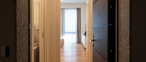 Un pasillo con suelo de madera, una puerta a la derecha y una ventana con cortinas al fondo.