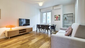 Una sala moderna con piso de madera, un sofá, un televisor, una mesa de comedor con sillas y una lámpara colgante.