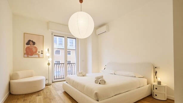 Un dormitorio con una cama grande, una silla, una lámpara colgante y una ventana con vistas a edificios.