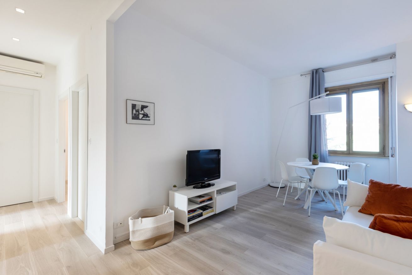 Una sala de estar moderna con un televisor de pantalla plana, muebles blancos y una zona de comedor con una mesa y sillas.