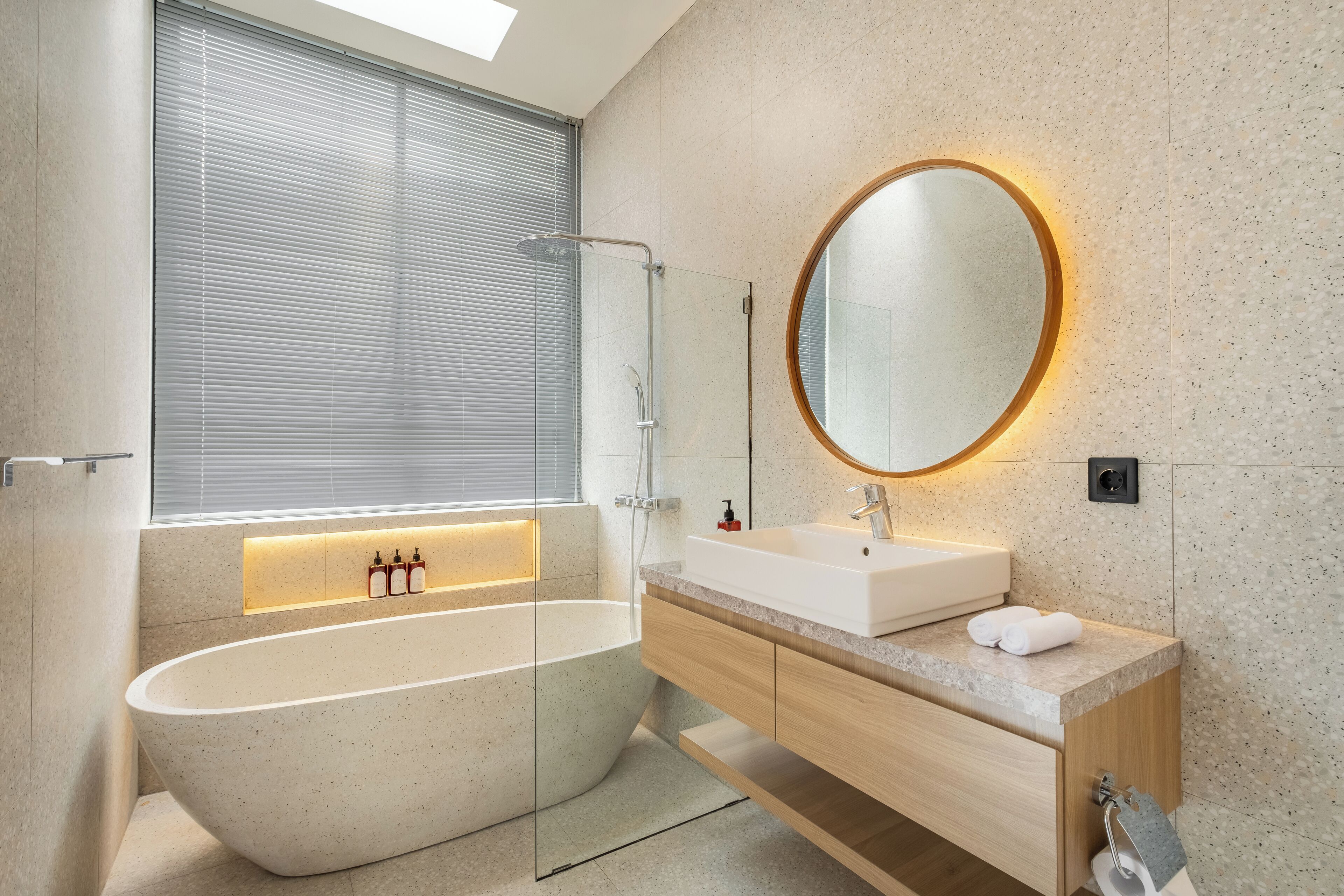 Un baño moderno con una bañera independiente, una ducha con cerramiento de vidrio, un tocador de madera con un lavabo blanco y un espejo grande con marco circular.