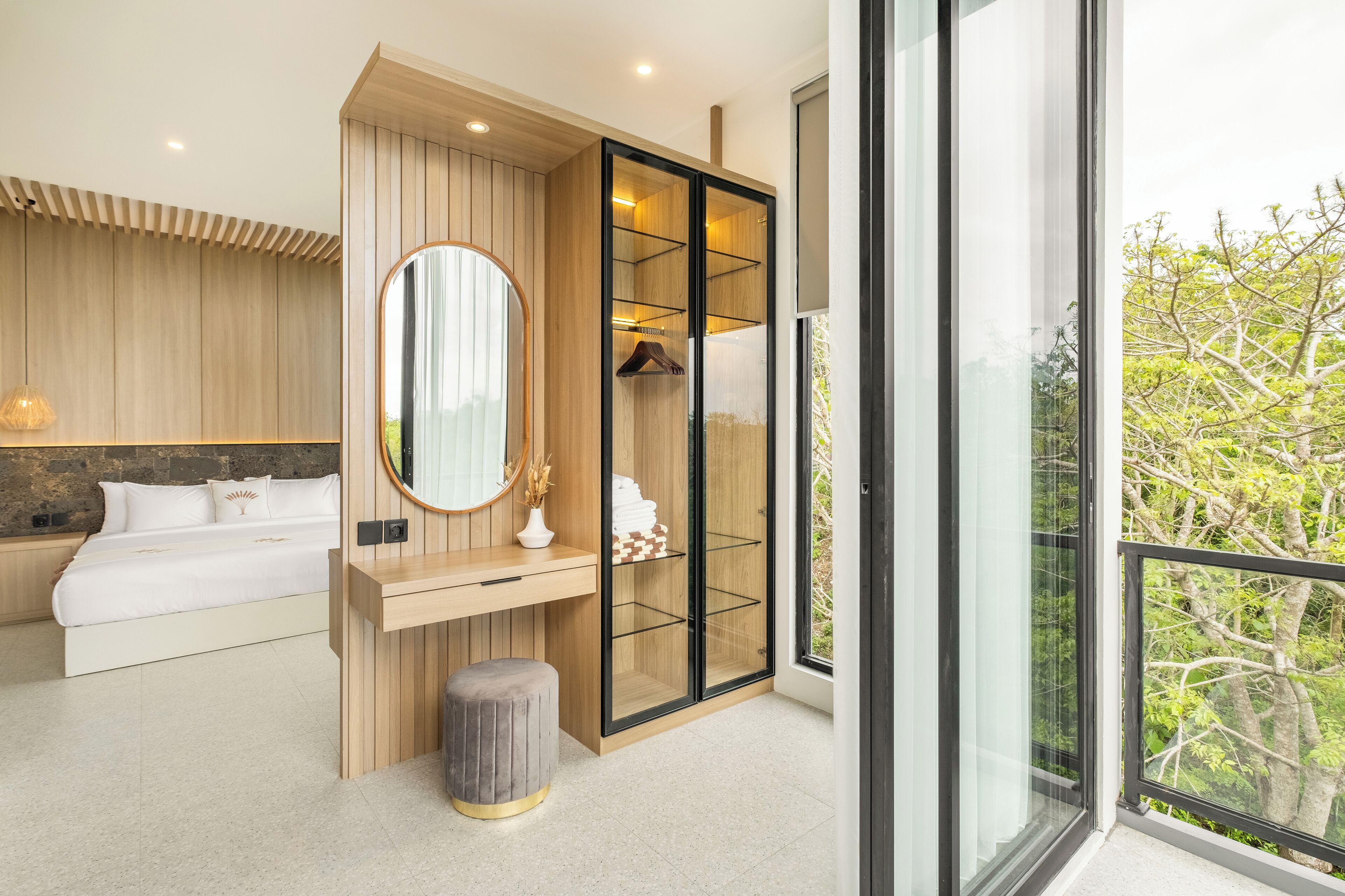 A modern bedroom with a bed, bedside table, mirror, wardrobe, and a view of trees through a large window.