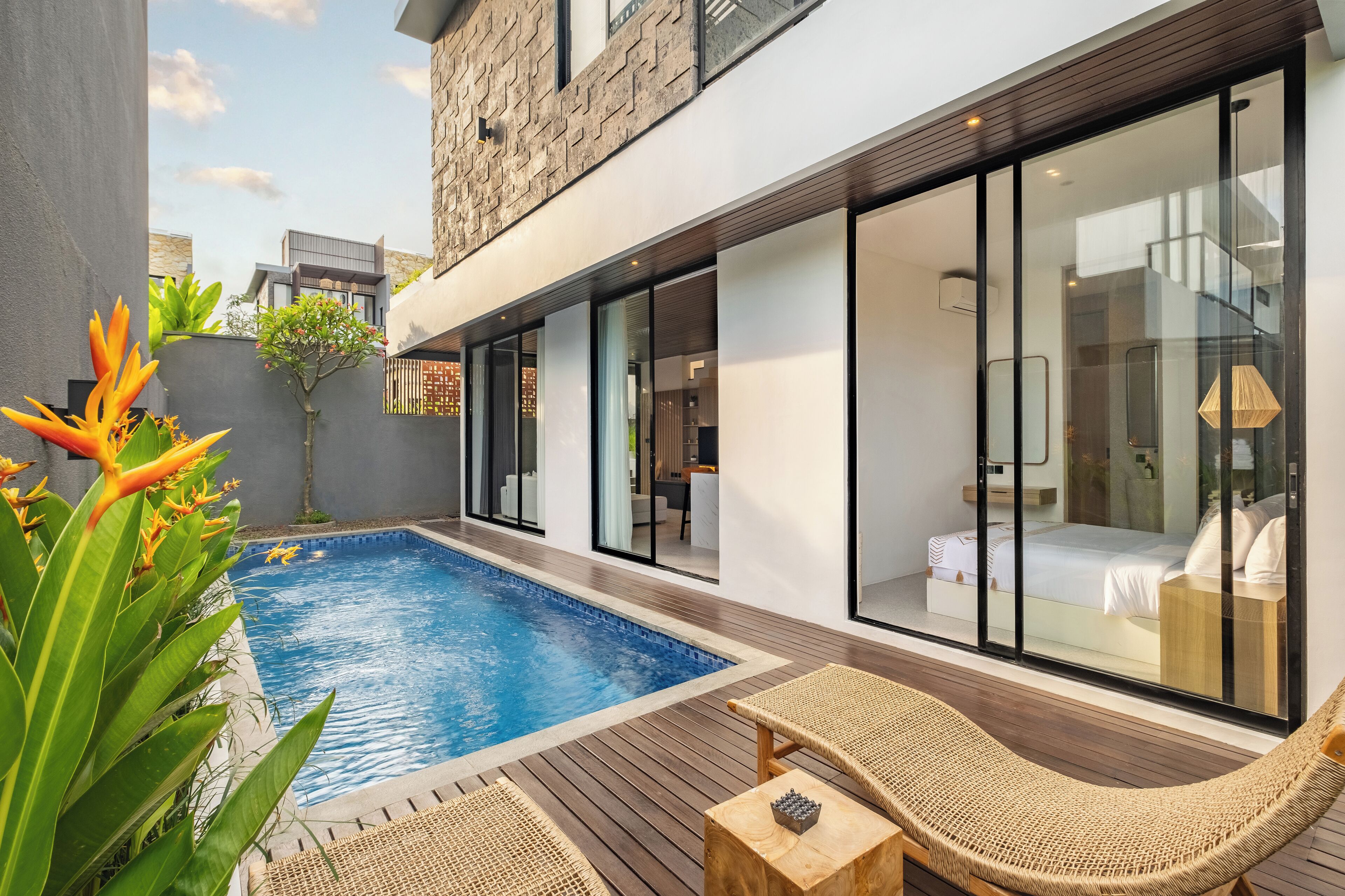 A modern poolside area with a wooden deck, lounge chairs, and a glass-enclosed bedroom with a bed and bedside table.