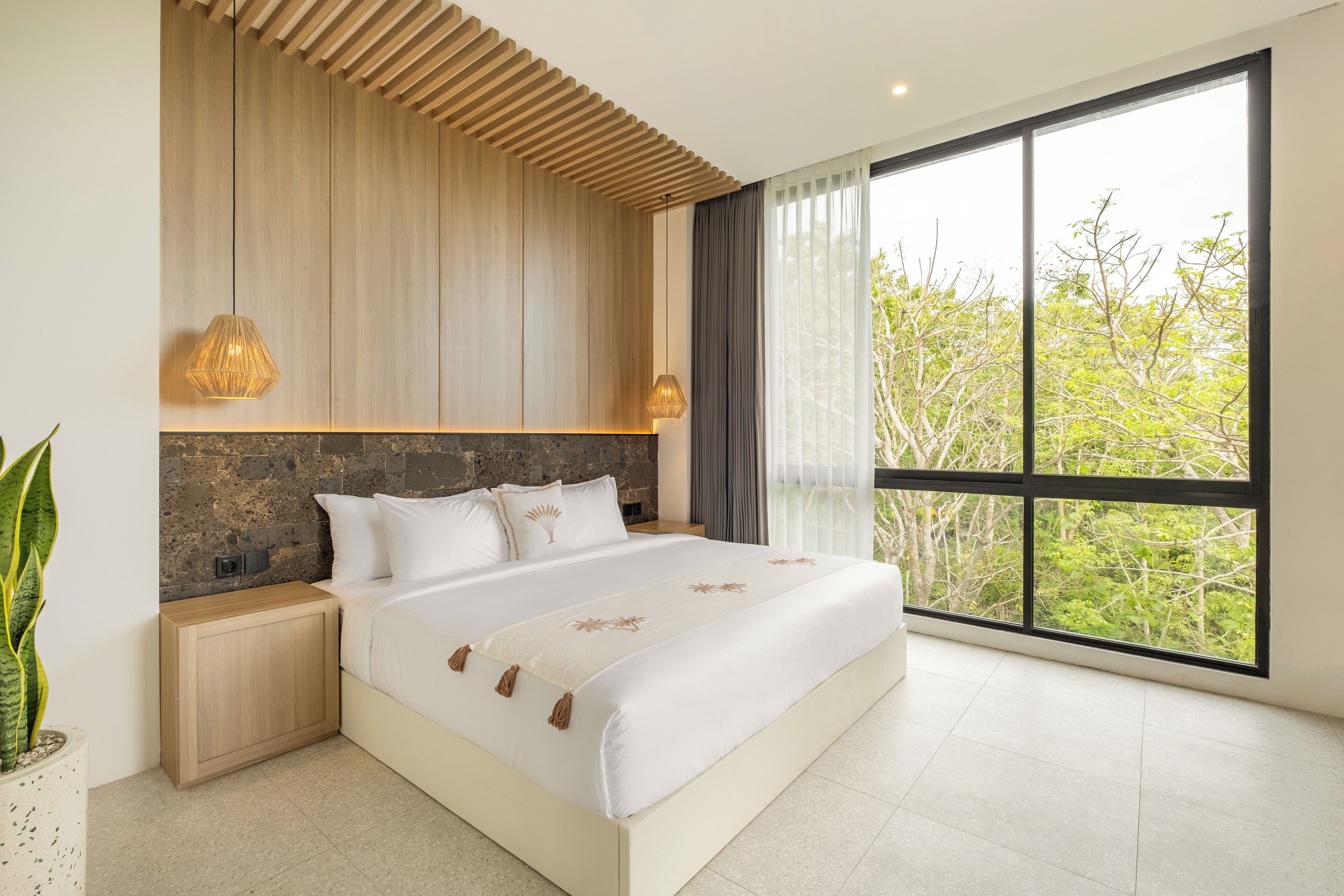 Une chambre à coucher moderne avec un grand lit, une tête de lit en bois et une vue sur les arbres à travers de grandes fenêtres.