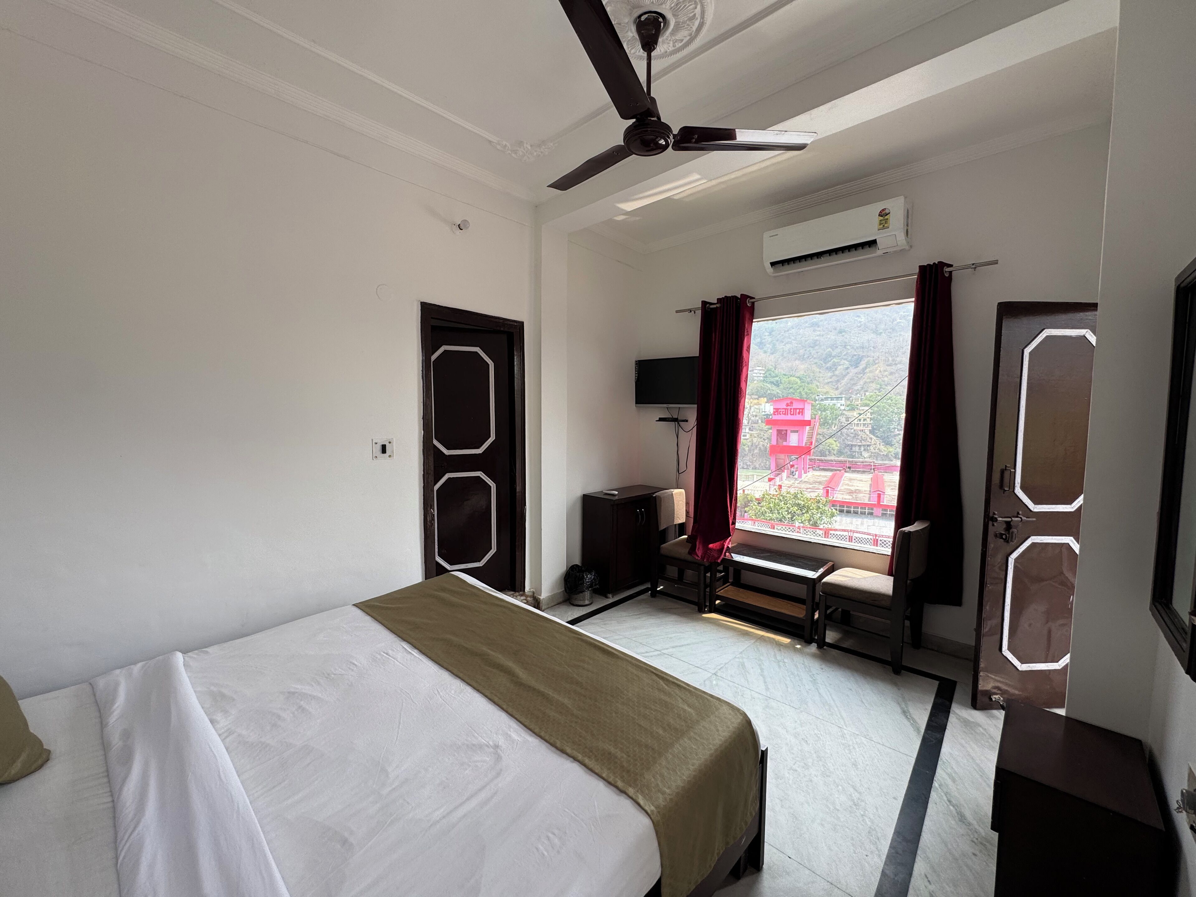 A hotel room with a bed, a desk, a chair, a ceiling fan, and a window with a view of a red building.