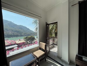 A room with a large window offering a view of a river and hills. There is a chair and a small table in the room. - Nirvana Riverside Resort (Rishikesh)