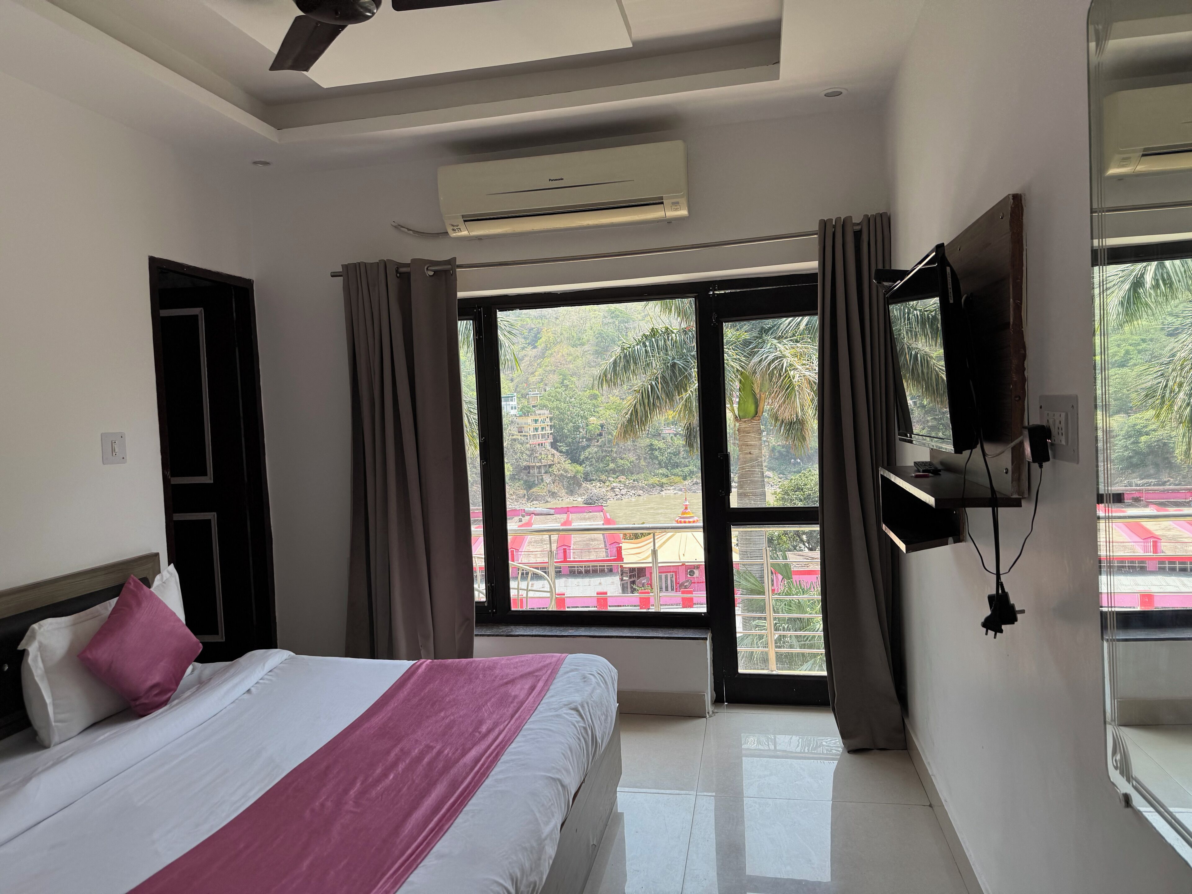 A hotel room with a bed, a TV mounted on the wall, and a large window offering a view of the outdoors.