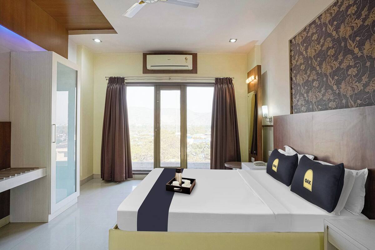 Ein Hotelzimmer mit einem großen Bett, einem Schreibtisch, einem Sessel, einem kleinen Tisch, einem Nachttisch, einem Spiegel, einem Fenster mit Vorhängen und Blick ins Freie.