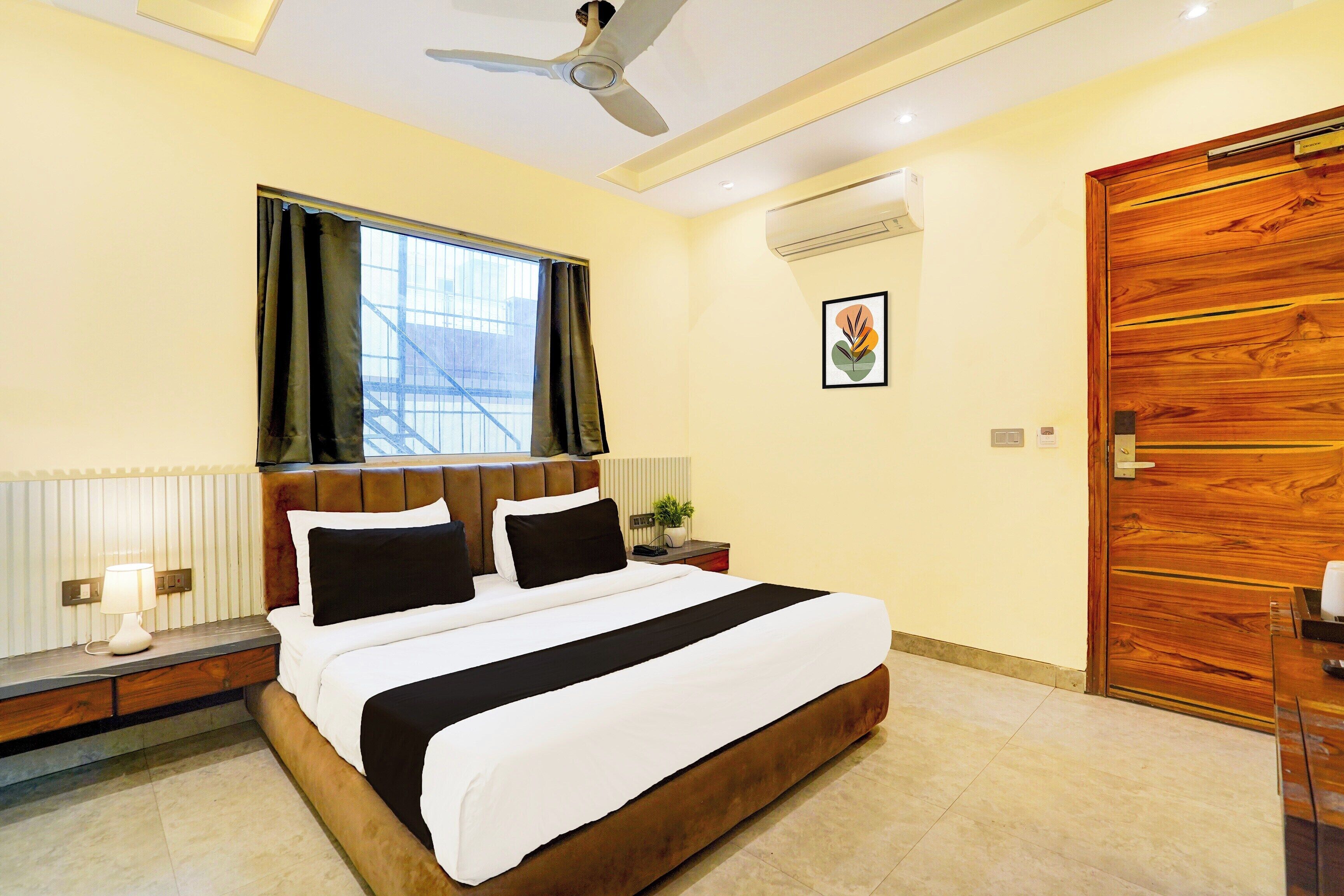 A bedroom with a large bed, a ceiling fan, a window with curtains, a wooden door, and a framed picture on the wall.