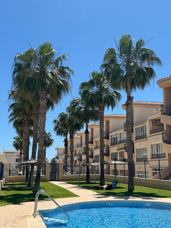 A pool area with palm trees, a clear sky, and a building with balconies.
