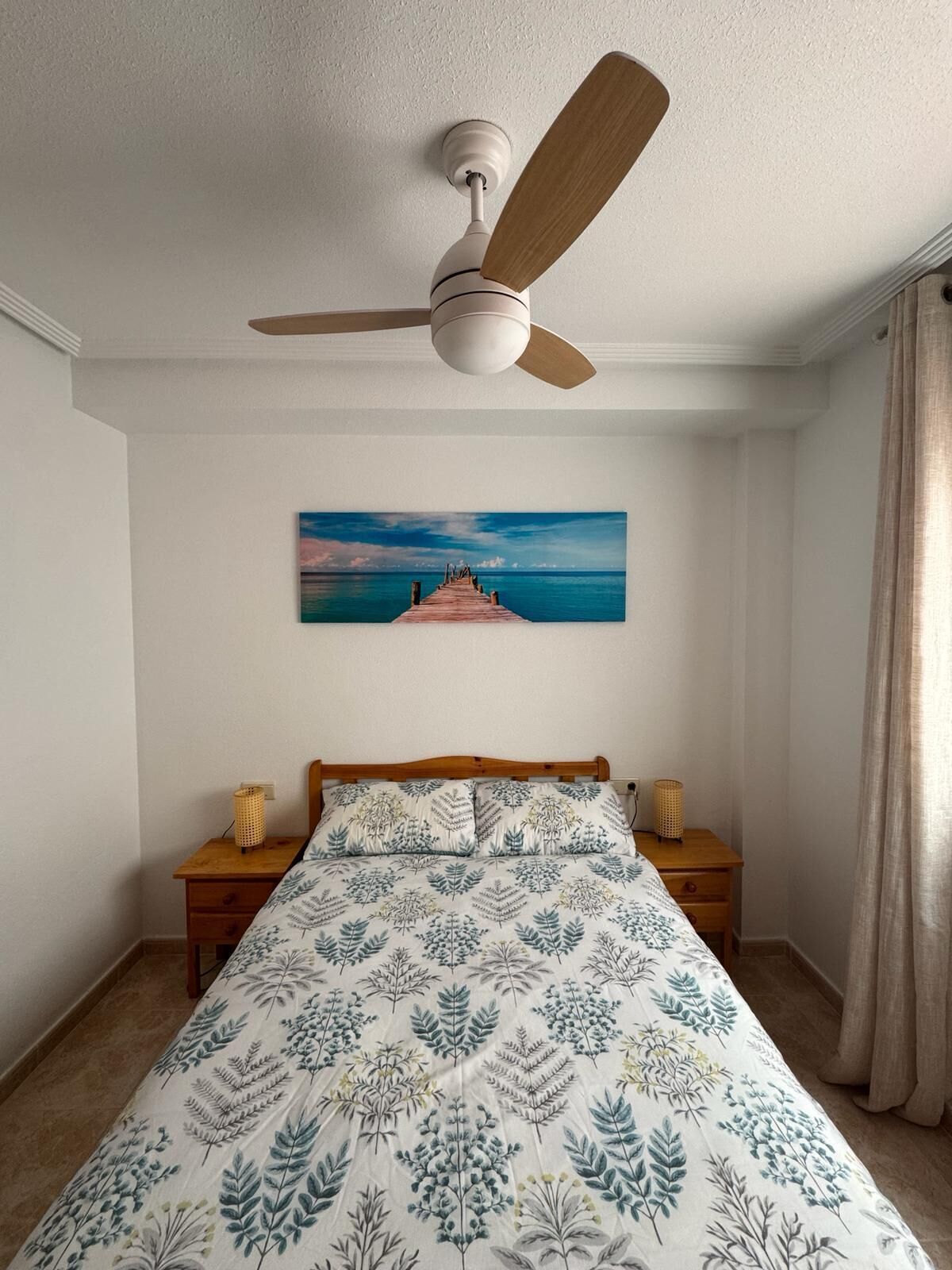 Une chambre à coucher avec un lit, un ventilateur de plafond, une table de chevet, une lampe, un tableau et une fenêtre avec des rideaux.