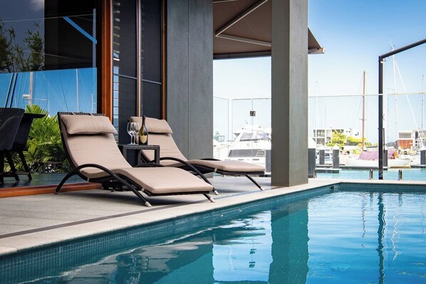 A pool area with lounge chairs and a table, a clear view of boats in the background, and a modern building facade.