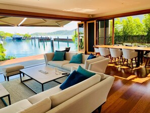 A modern living room with a large window overlooking a body of water, a dining area with a wooden table and chairs, and a sofa set with cushions.