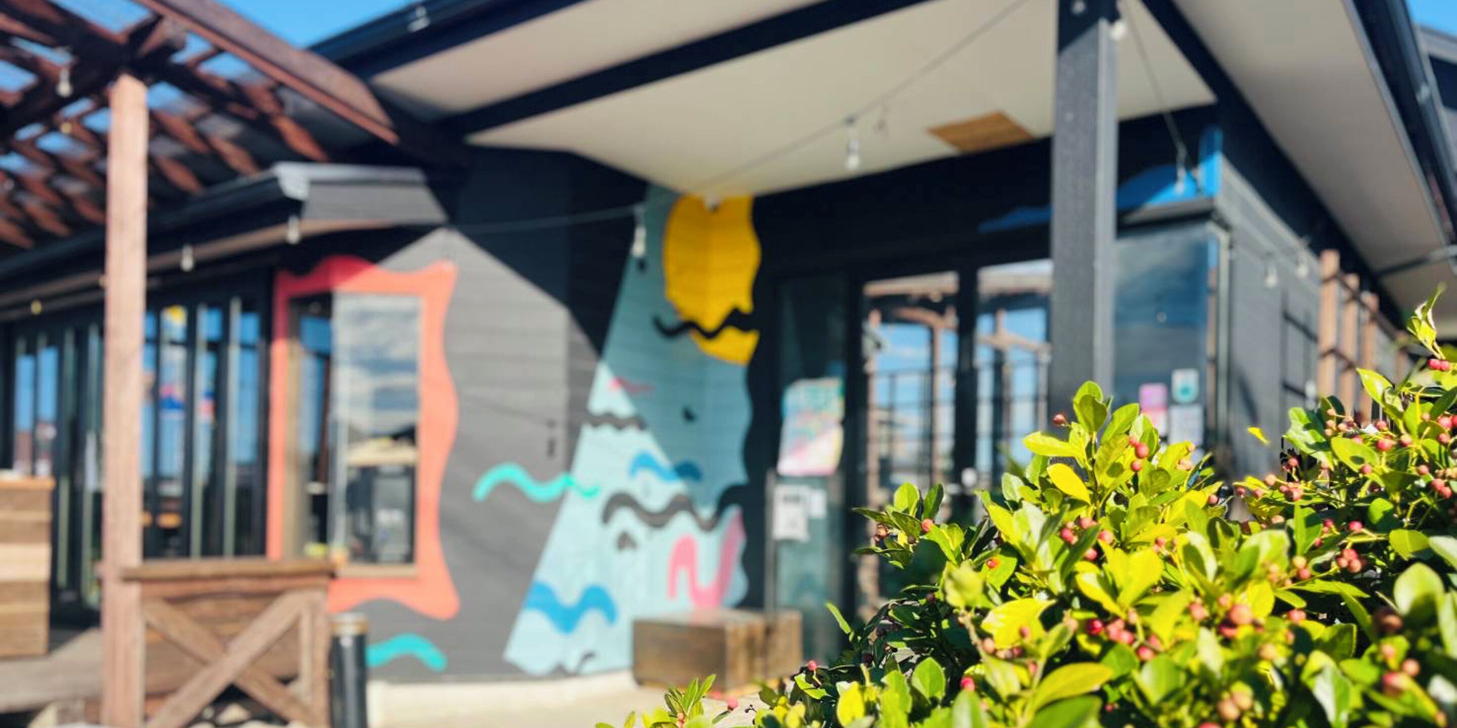 A modern building with a colorful mural on the exterior wall, a covered outdoor area, and lush greenery in the foreground.