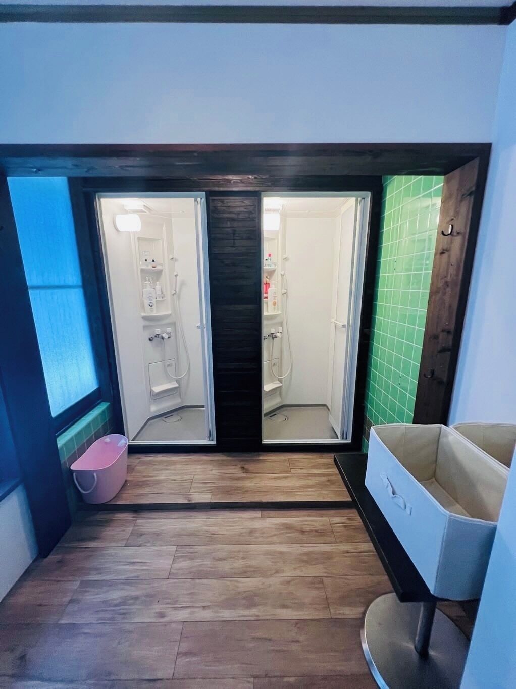 Un couloir étroit, doté d’un plancher en bois, mène à une salle de bain équipée d’une douche, de toilettes et d’un lavabo. On y trouve une petite poubelle rose et une chaise blanche.