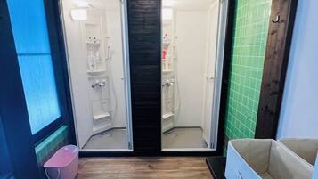 En smal gang med trægulv, der fører til et badeværelse med bruser, toilet og håndvask. Der er en lille pink skraldespand og en hvid stol i gangen.