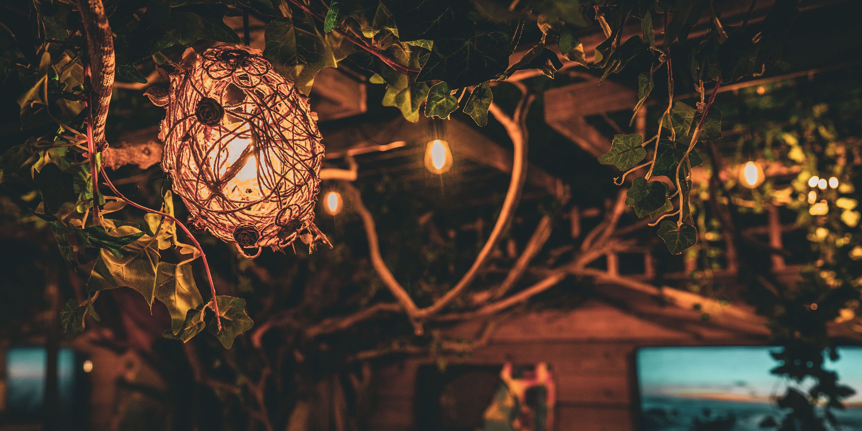 Un luminaire suspendu, composé de branches entrelacées, diffusant une lumière chaleureuse, le tout mis en valeur par une structure en bois et un décor de feuillage.