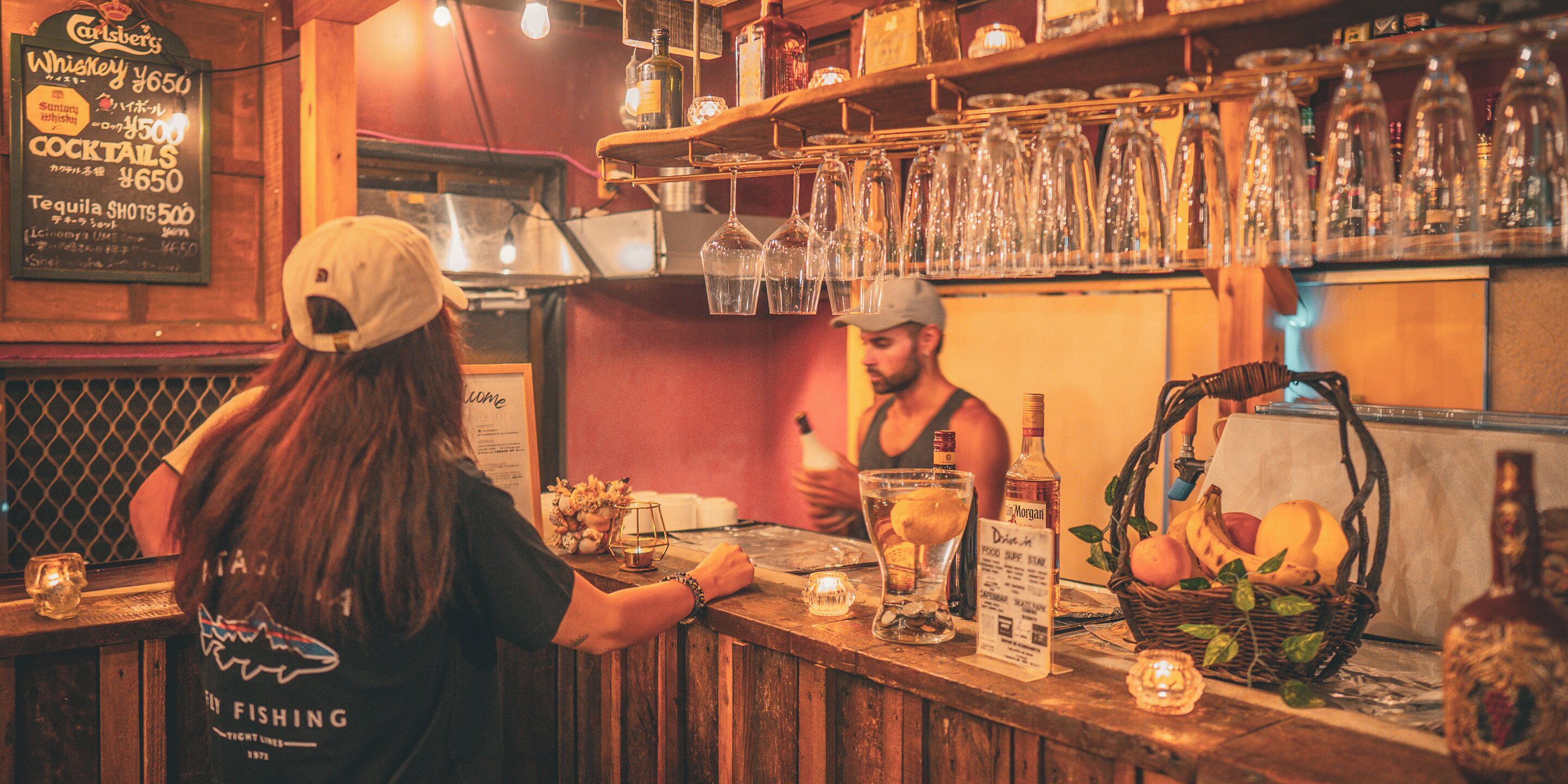 Uma área de bar com um cardápio, copos, garrafas e uma cesta de frutas.