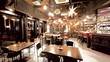 Zona de bar con mesas y sillas, una barra con un barista, y una variedad de botellas y vasos en estanterías.