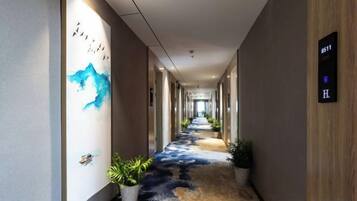 Un couloir d’hôtel avec un sol recouvert de moquette, un mur orné d’un tableau abstrait bleu et un panneau indiquant le numéro de chambre : 8511.