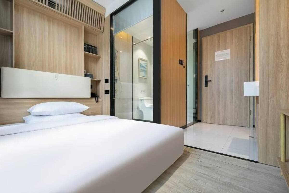 A modern hotel room with a large bed, wooden paneling, a glass shower enclosure, and a wooden door.