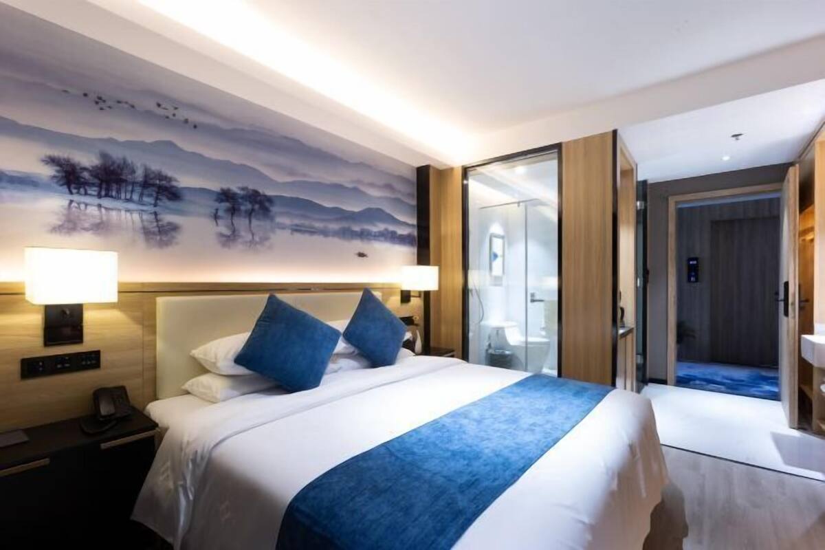 A hotel room with a bed, two bedside lamps, a nightstand, a wall mural, and a bathroom door.